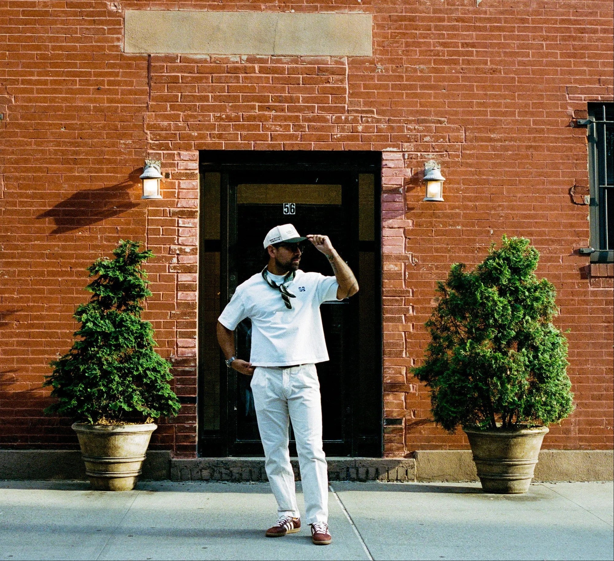 A man wearing a white polo shirt, white trousers, and a white cap stands in front of a red brick building doorway, with two potted green trees on either side.