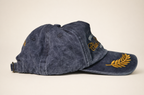 A washed navy blue baseball cap with gold embroidered details, including a fern leaf on the brim and script embroidery on the side, shown against a plain beige background.