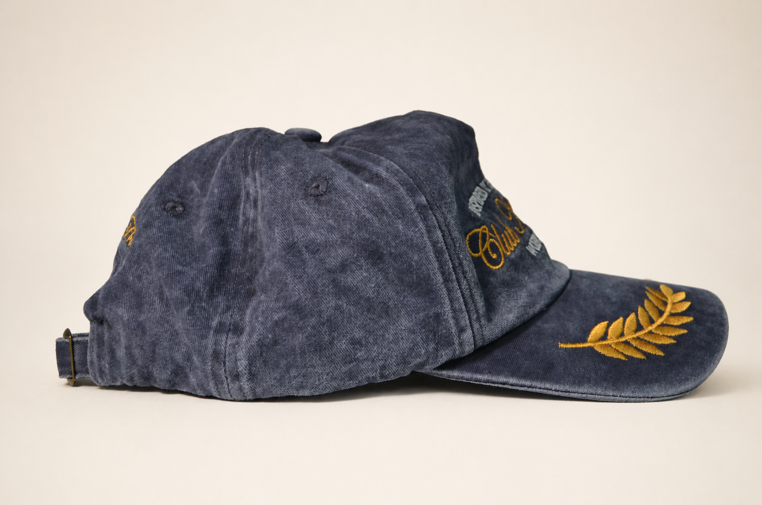 A washed navy blue baseball cap with gold embroidered details, including a fern leaf on the brim and script embroidery on the side, shown against a plain beige background.