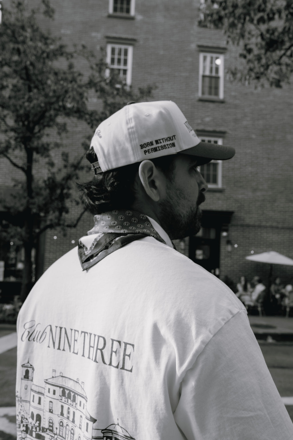 A man in a white 'Don't Tread on Me' baseball cap and a white 'Eliot Ninethree' graphic t-shirt, photographed from behind in an outdoor urban setting.