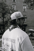 A man in a white 'Don't Tread on Me' baseball cap and a white 'Eliot Ninethree' graphic t-shirt, photographed from behind in an outdoor urban setting.