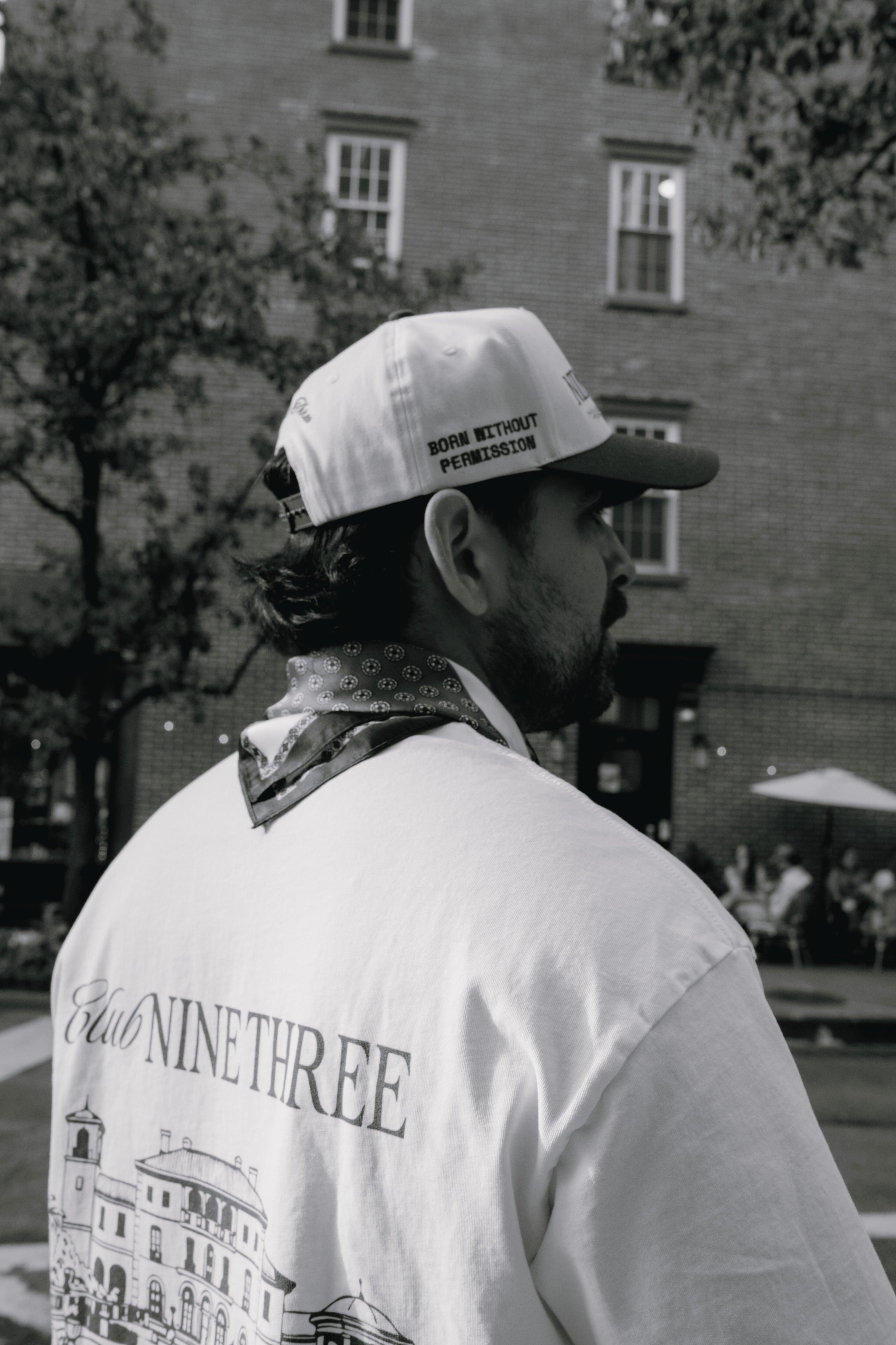A man in a white 'Don't Tread on Me' baseball cap and a white 'Eliot Ninethree' graphic t-shirt, photographed from behind in an outdoor urban setting.